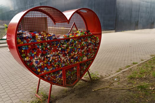 A heart-shaped metal container filled with colorful plastic plugs stands in a city park, promoting recycling efforts and environmental awareness.