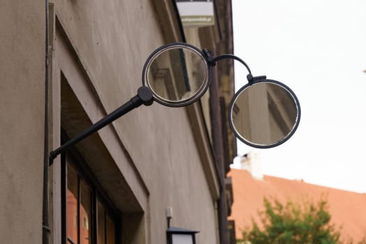 A vintage glasses shop sign featuring a mirrored glasses frame is mounted on the exterior wall of a building, advertising the shops location. The sign is positioned on a street with other buildings visible in the background.