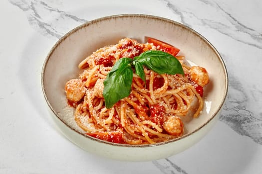 Bowl of Margherite style spaghetti with rich tomato sauce, juicy meatballs and sprinkle of grated parmesan, garnished with leaves of fresh aromatic basil, presented on marble surface