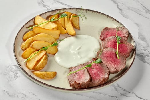 Tender juicy slices of roast beef served with golden baked potato wedges and smooth cream sauce, garnished with fresh green pea shoots on marble table
