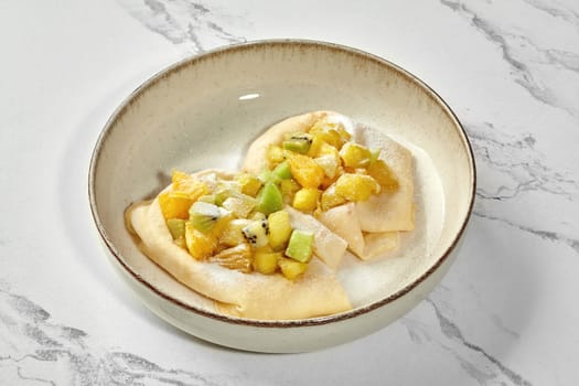 Delicate sweet thin homemade crepes topped with diced fresh fruit and dusting of powdered sugar, served in ceramic bowl against marble background. Nutritious breakfast for indulgent start to day