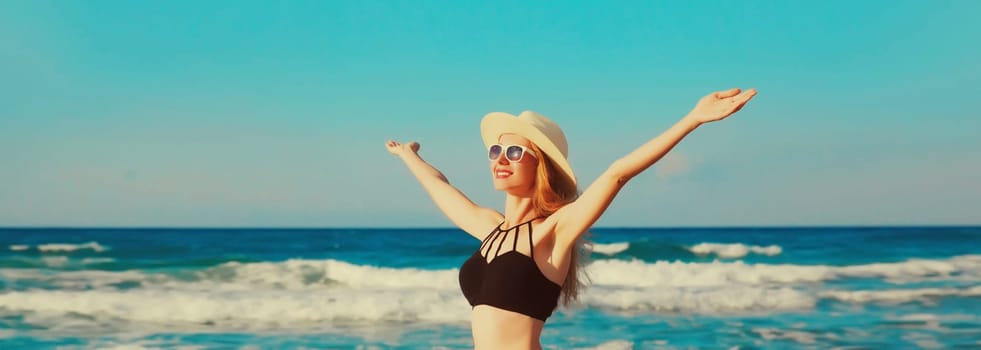 Summer vacation, happy smiling woman in bikini swimsuit and straw hat raising her hands up on the beach on sea coast background