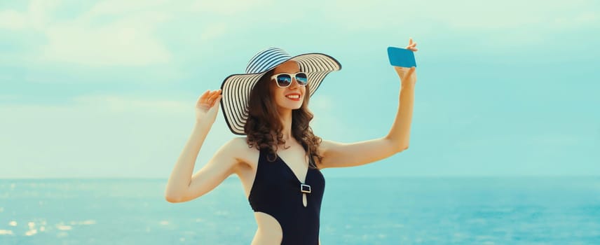 Summer portrait of beautiful young woman taking selfie with smartphone on the beach wearing straw hat and bikini on sea background