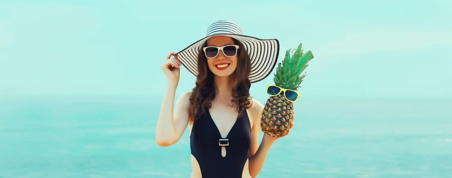 Summer vacation, beautiful happy smiling woman in black bikini swimsuit and straw hat holding pineapple fruits on the beach on sea coast background