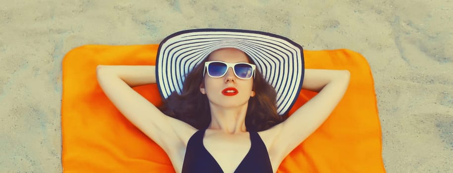Summer vacation, portrait of beautiful young woman lying on sand on the beach wearing a black bikini swimsuit and straw hat