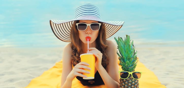 Summer vacation, happy relaxed woman drinking juice with pineapple in straw hat lying on sand on the beach on sea background