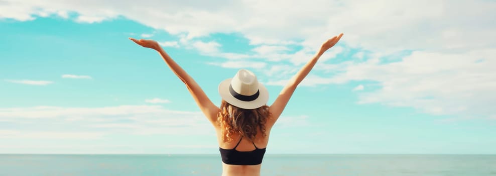 Summer vacation, rear view of beautiful happy slim woman in bikini swimsuit and straw hat raising her hands up on the beach on sunny day on sea coast background