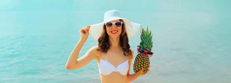 Summer vacation, beautiful happy smiling woman in bikini swimsuit and straw hat holding pineapple fruits on the beach on sea coast background on sunny day