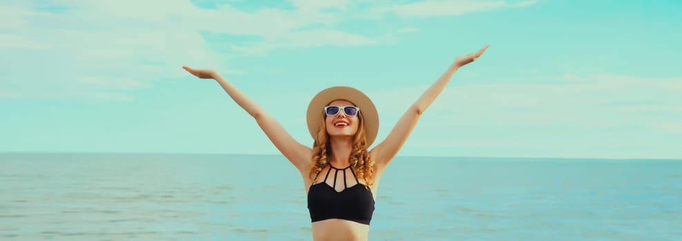 Summer vacation, happy smiling woman in bikini swimsuit and straw hat raising her hands up on the beach on sea coast background