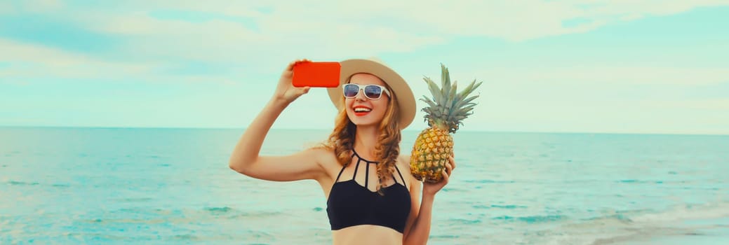 Summer vacation, happy smiling young woman holding pineapple taking selfie with smartphone wearing bikini swimsuit and straw hat on the beach on sea coast background