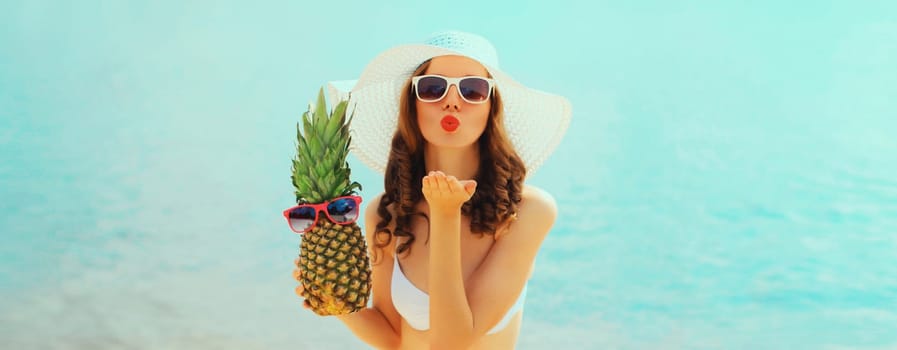 Summer vacation, beautiful happy young woman in bikini swimsuit and straw hat holding pineapple fruits blowing her lips sends kiss on the beach on sea coast background on sunny day