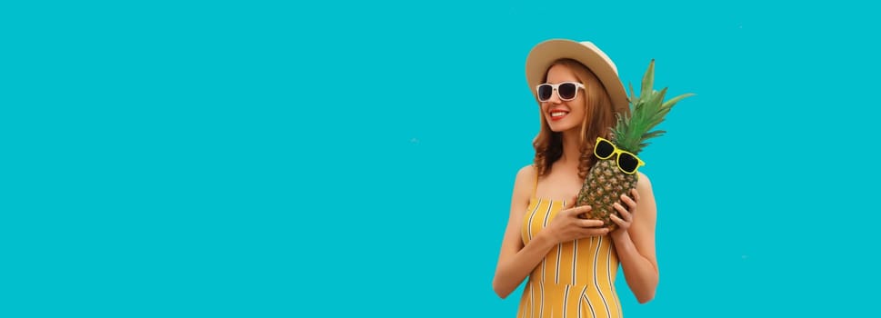 Summer portrait of happy smiling young woman with pineapple fruit in straw tourist hat, sunglasses on blue studio background