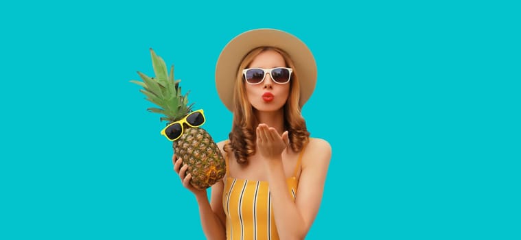 Summer portrait of beautiful young woman blowing a kiss with pineapple wearing straw hat, glasses posing on blue studio background