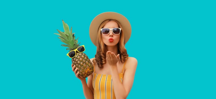 Summer portrait of beautiful young woman blowing a kiss with pineapple wearing straw hat, glasses posing on blue studio background