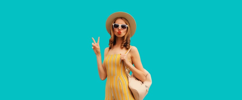 Beautiful blonde young woman blowing a kiss posing wearing summer hat, backpack on blue studio background