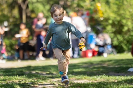 A young boy is running through a park with a toy bus in his hand. The scene is lively and playful, with other children and adults in the background. The boy's excitement and energy are contagious