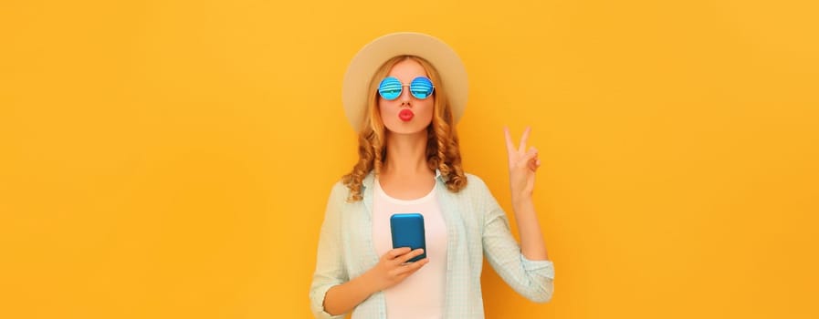 Portrait of stylish young woman posing blowing a kiss with smartphone wearing summer straw hat posing on yellow background