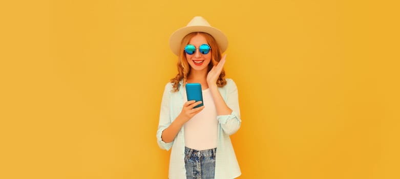 Summer portrait of happy surprised amazed young woman with mobile phone looking at device in straw tourist hat on yellow studio background