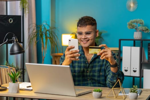 Happy Indian man using bank credit card smartphone for online shopping payments at home office. Business transaction. Smiling guy freelancer purchases with cellphone E-banking app service. Lifestyle