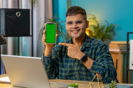 Indian young businessman holding smartphone with green screen chroma key mock up recommend good application promotional sale offer. Freelancer man guy at home office desk looking at camera. Lifestyle