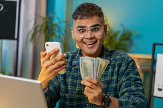 Planning family budget. Amazed happy Indian man counting money cash use smartphone calculate domestic bills. Hispanic guy satisfied of income and saves salary money at home office table workplace