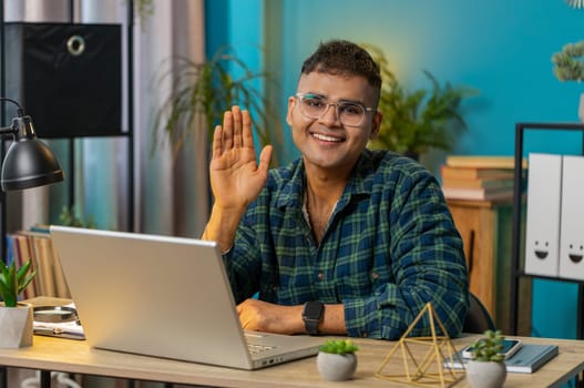 Hello. Indian man smiling friendly at camera and waving hands gesturing hello, hi, greeting or goodbye welcoming, online video call POV conversation. Guy at home office table workplace. Lifestyle