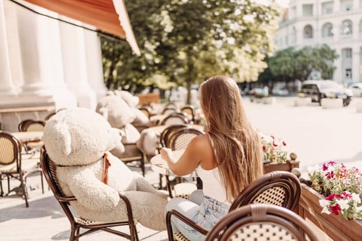 A woman sits cafe with a teddy bear next to her. The scene is set in a city with several chairs and tables around her. The woman is enjoying her time at the outdoor cafe