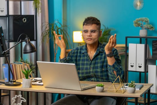 Indian man working on laptop, breathes deeply, eyes closed meditating with concentrated thoughts, peaceful mind. Hispanic guy taking a break, relaxing sitting at home office table workplace. Lifestyle