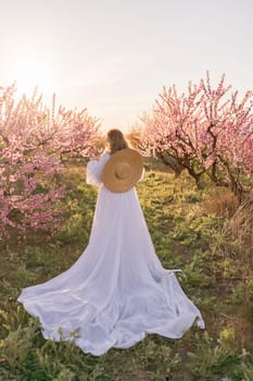 Woman blooming peach orchard. Against the backdrop of a picturesque peach orchard, a woman in a long white dress and hat enjoys a peaceful walk in the park, surrounded by the beauty of nature