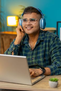 Happy Hispanic man sits at home office desk listening to favorite energetic disco music through wireless headphones. Hindu freelancer guy relaxing taking a break with laptop on table. Vertical shot