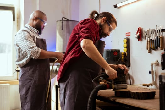 Carpenter collaborating with african american apprentice using orbital sander with fine sandpaper to achieve refined finish. Man and coworker using angle grinder on wood block for professional results