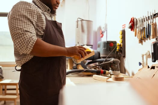 African american man in assembly shop using sandpaper for sanding wooden surface before painting it, ensuring adequate finish. BIPOC carpenter using abrasive sponge to repair damages on wood block