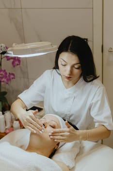 A young beautiful Caucasian brunette girl cosmetologist smears cleansing cream with both hands on the face of an adult female client, which lies with her eyes closed on a massage table in a beauty salon, side view close-up.