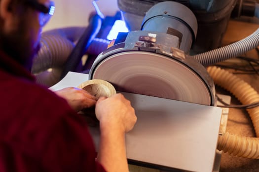 Artisan sanding wooden object on disc sander in assembling shop. Diy hobby enjoyer working in joinery, guiding wood against grinding machine rotating disc to create art