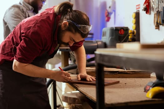 Craftsperson using ruler to take measurements of wood plank with coworker in background solving tasks. Man drawing with pencil on wooden board to determine where to make cut near BIPOC colleague