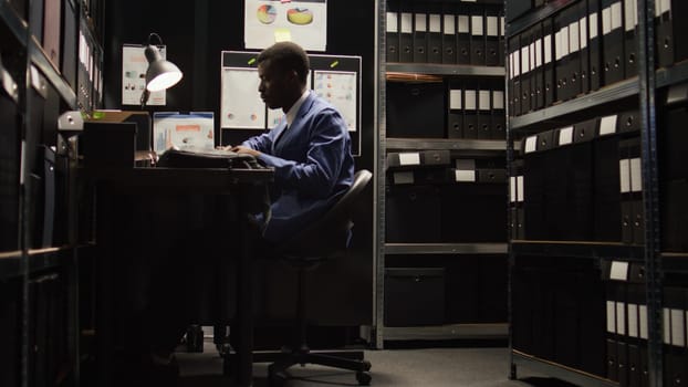 Young law officer with experience in criminal investigations enters the office and turns on his laptop. African American investigator starts researching while seated in workroom with laptop bag.