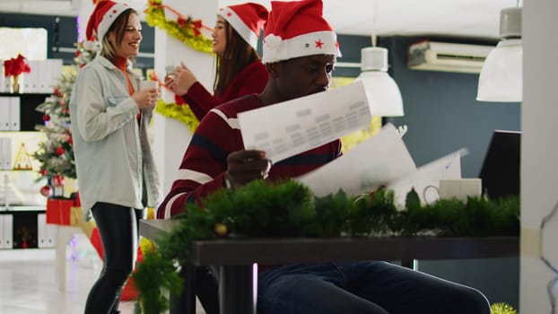 African american employee in Christmas decorated office working on overdue project while carefree coworkers enjoy coffee break together. Worker frustrated by women laughing and distracting him