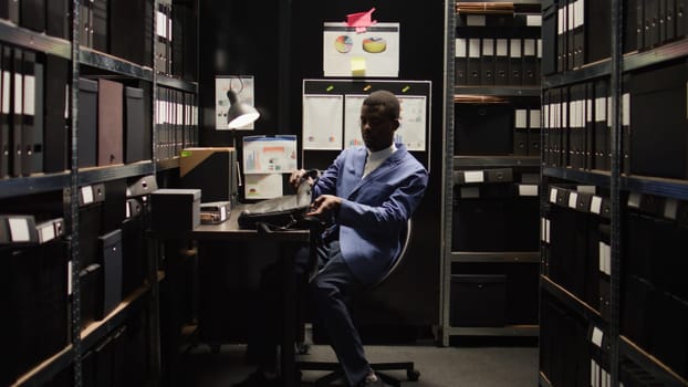 Male detective inspector carefully examines evidence files while setting personal computer down on table. African american police officer entering archive room with laptop bag, ready to solve crimes.