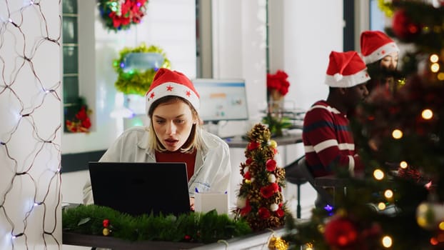 Focused woman in Christmas ornate office looking at computer screen, paying attention. Diligent employee checking emails in multiethnic xmas adorn workplace during festive holiday season