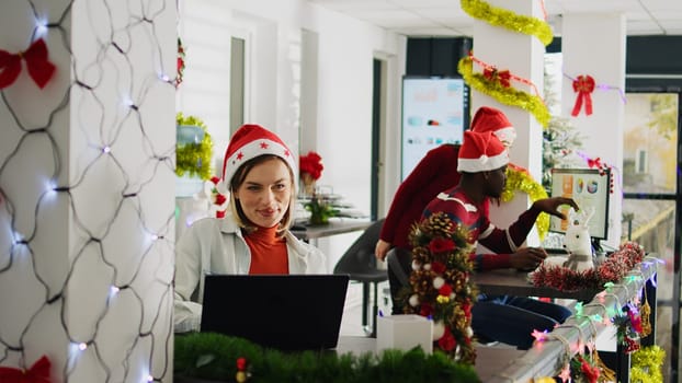 Happy multiethnic team working on christmas season in decorated office. Cheerful employees solving tasks at desk in diverse workplace next to colleagues during winter festive time