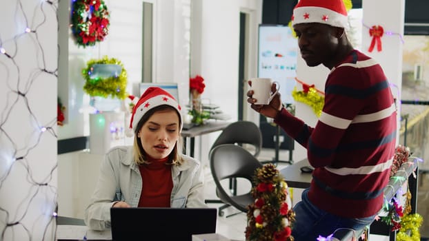 Zoom in shot of african american employee taking a break, enjoying coffee in Christmas decorated office. Staff member discussing with colleague wearing Santa hat during festive holiday season