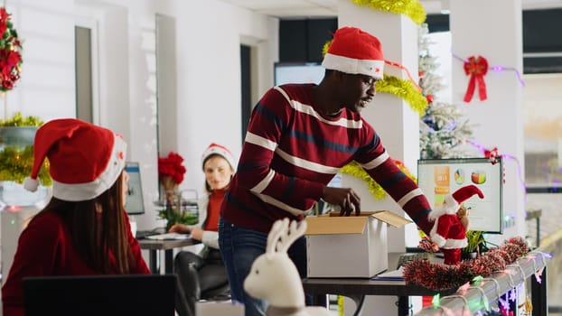 Happy african american employee clearing festive decorated office desk before leaving for vacation. Worker wearing Santa hat taking days off to go on xmas holiday, feeling joyful