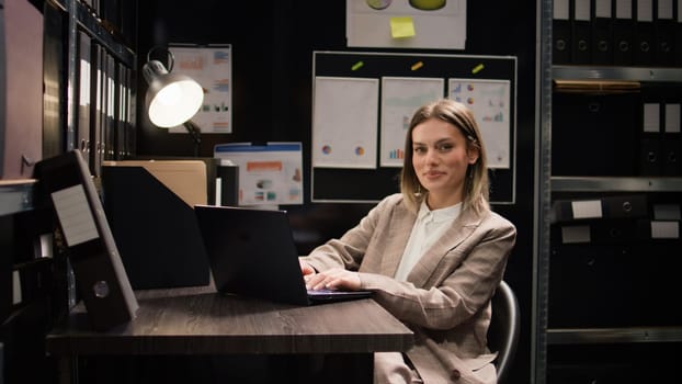 Private investigator examines evidence, looks through documents, and uses laptop in office. Portrait view of female police officer searching criminal records and files on minicomputer. Zoom-out shot.