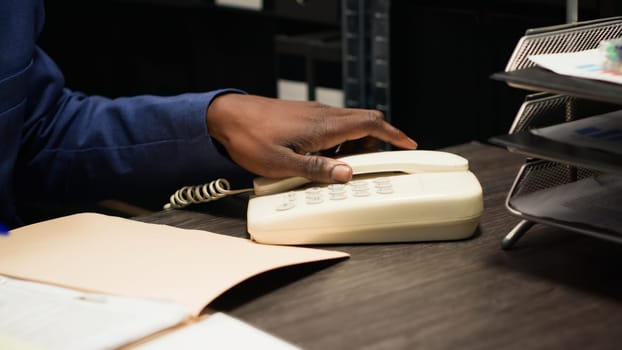 Detailed shot of african american officer answering call on landline and takes crucial notes of case. Expert male detective uses telephone to gather evidence and analyze clues in well-equipped office.