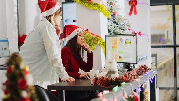 Employee wearing Santa hat working in Christmas ornate office requesting help from coworker. Staff member looking over business revenue numbers with colleague in xmas adorn workplace, handheld camera