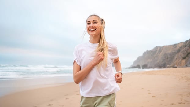 Smiling young woman runner with blond hair in white top jogging on sandy beach. Cheerful female athlete jogging along sea under sky. Happy girl represents healthy lifestyle.
