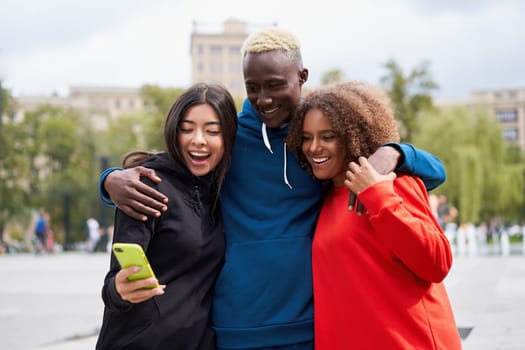 Cheerful diverse young man with female friends standing arm around looking at smartphone screen in city. Happy students of different nationalities spending weekend time together in city park.