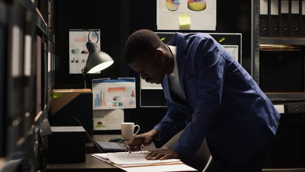 Private investigator grabbing files, analyzing statements, and using laptop to unveil truth in investigation. African american officer utilizing personal computer to cross check with evidence papers.