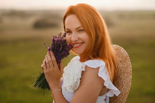 A girl in a white dress with a bouquet of lavender flowers stands in a field at sunset.