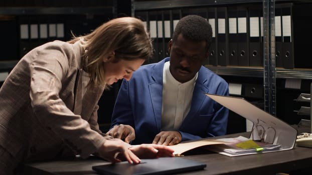 African american detective holding case files enters office with caucasian policewoman carrying backpack. Policeman discussing clues and evidence while female investigator places laptop on desk.